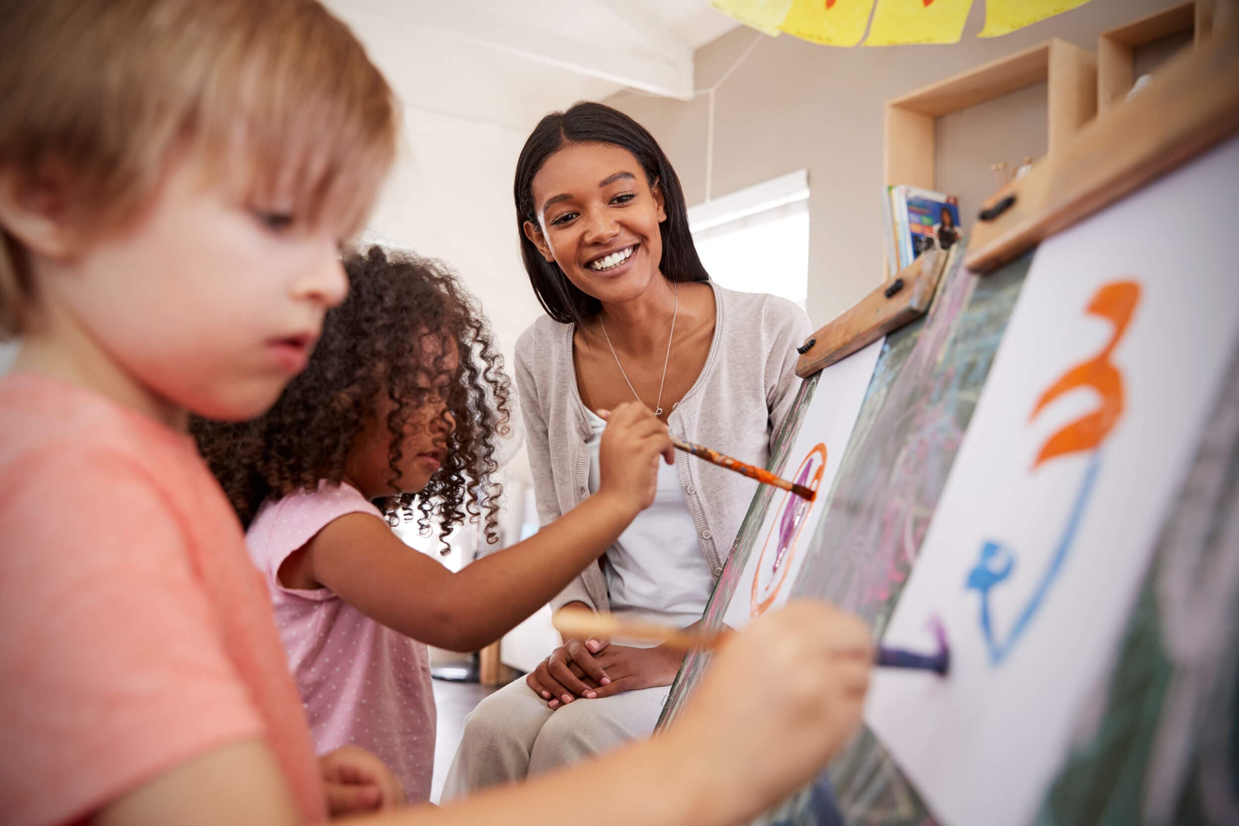 A teacher painting at an easel with two young children