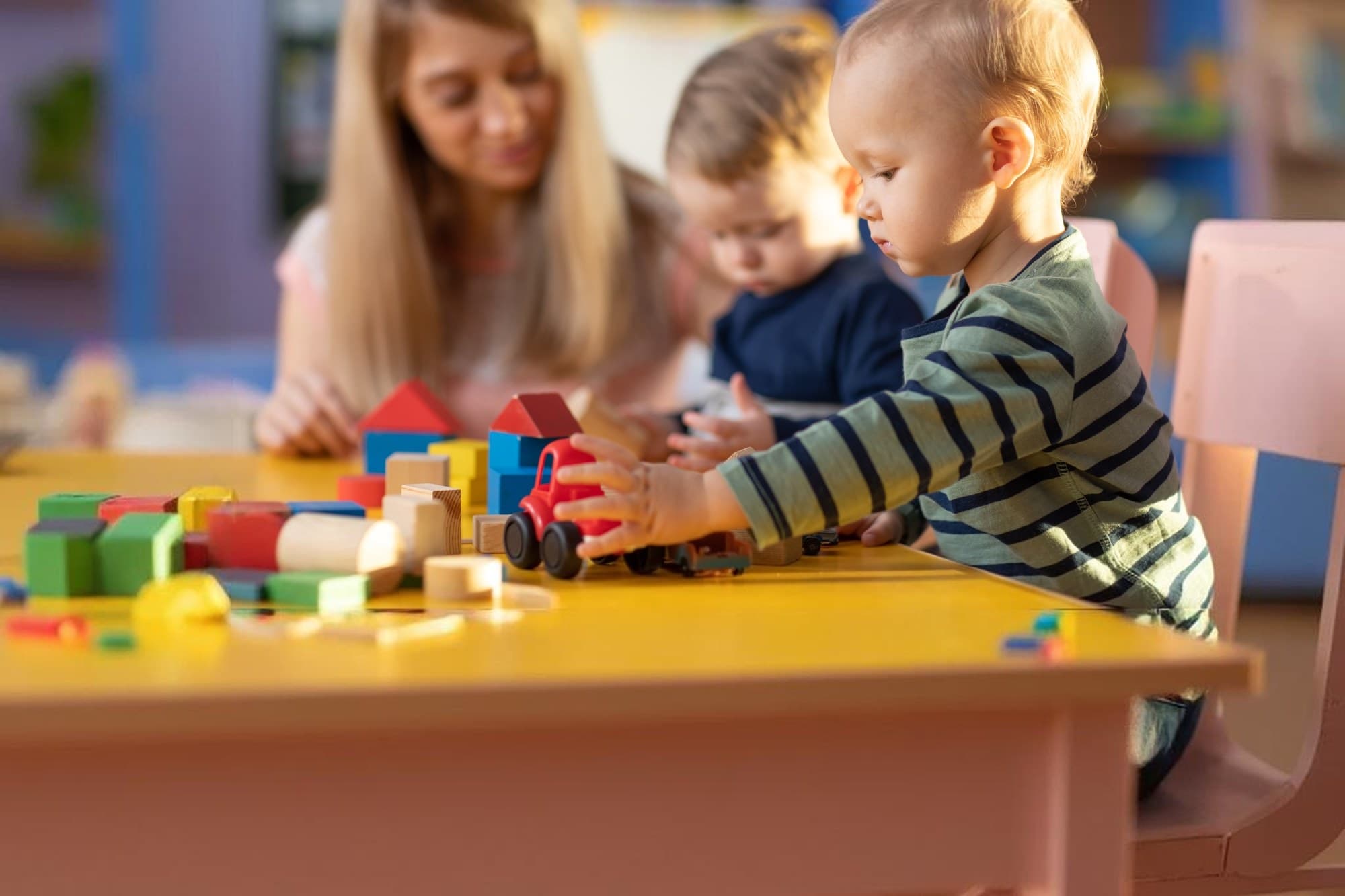 A teacher and two toddlers playing with wooden blocks and toy cars at a classroom table